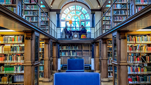 Interior of a historic library with tall wooden bookshelves filled with books, ornate vaulted ceiling with decorative patterns, a large arched window at the back, and a blue upholstered chair in the centre on a parquet floor.
