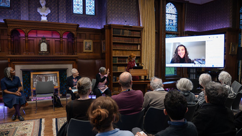 Audience attending a prize-giving ceremony in Girton College’s wood-paneled library, with speakers seated at the front and a large screen for remote participation.