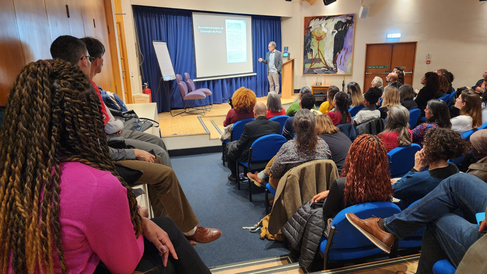 Audience listen as Professor João José Reis presents the lecture from a small stage in front of a powerpoint slide