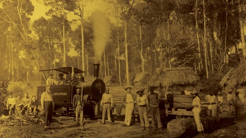 Archival image of a steam locomotive labeled “Enterprise” with smoke rising from its chimney, positioned on rail tracks in a forest clearing. Several workers stand around the train and timber logs, with thatched huts visible among tall trees in the background.