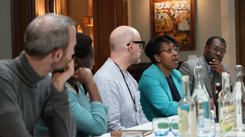 Group of people seated around a table in a meeting room, with water bottles and glasses on the table. The background shows a wooden door and a framed painting on the wall.