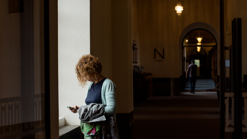A person standing by a window inside a quiet university hallway, looking down at something in their hands, with warm hanging lights illuminating the long corridor.