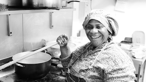 A person working in a kitchen stands beside a stove, stirring a large pot with a long wooden utensil. The setting includes metal cookware, a tiled wall, and various kitchen equipment in the background.