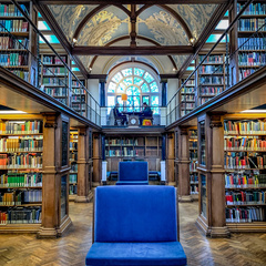 Interior of a historic library with tall wooden bookshelves filled with books, ornate vaulted ceiling with decorative patterns, a large arched window at the back, and a blue upholstered chair in the centre on a parquet floor.
