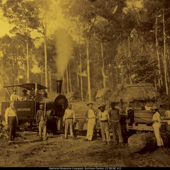 Archival image of a steam locomotive labeled “Enterprise” with smoke rising from its chimney, positioned on rail tracks in a forest clearing. Several workers stand around the train and timber logs, with thatched huts visible among tall trees in the background.