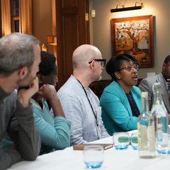 Group of people seated around a table in a meeting room, with water bottles and glasses on the table. The background shows a wooden door and a framed painting on the wall.