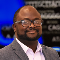 Portrait photo of Dr Jerome Atutornu, smiling and looking at the camera, wearing glasses and a dark suit jacket over a light shirt, with a blurred indoor background.