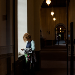 A person standing by a window inside a quiet university hallway, looking down at something in their hands, with warm hanging lights illuminating the long corridor.