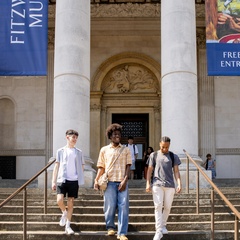 Three University of Cambridge students walk down the stairs in front of tall white pillars and banners outside the Fitzwilliam Museum.