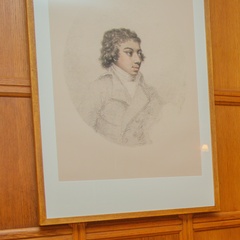 A framed pencil drawing hangs on a wooden-paneled wall. The sketch shows an adolescent figure in period clothing, with softly rendered shading and a loosely sketched jacket. The artwork is mounted within a wide mat and a simple wooden frame.