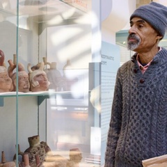 A person wearing a knitted sweater and hat stands inside a museum gallery, holding a book while looking at a glass display case