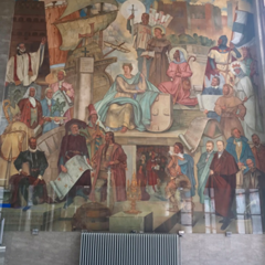A large wall mural depicting many historical scenes and figures arranged in tiers, including people holding books, maps and objects, with architectural elements and symbolic imagery throughout. A radiator sits below the mural.