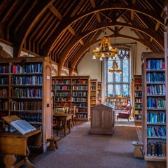 A view of a Girton College library with tall wooden bookshelves, a high timber‑framed ceiling, study tables, and a large arched window at the far end letting light into the room.