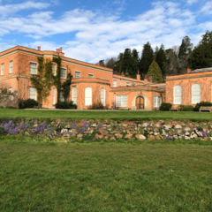A wide view of a peach‑coloured historic building with arched windows and ivy‑covered walls, set behind a green lawn and low stone wall, with trees and a blue, partly cloudy sky in the background.