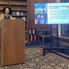 Dr. Tara Inniss speaking at a lectern during a presentation, with a slide on Barbadian rum-making displayed on a screen beside her.