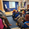 Audience listen as Professor João José Reis presents the lecture from a small stage in front of a powerpoint slide