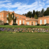A wide view of a peach‑coloured historic building with arched windows and ivy‑covered walls, set behind a green lawn and low stone wall, with trees and a blue, partly cloudy sky in the background.