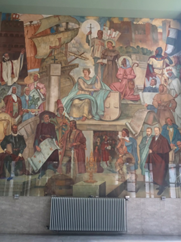 A large wall mural depicting many historical scenes and figures arranged in tiers, including people holding books, maps and objects, with architectural elements and symbolic imagery throughout. A radiator sits below the mural.