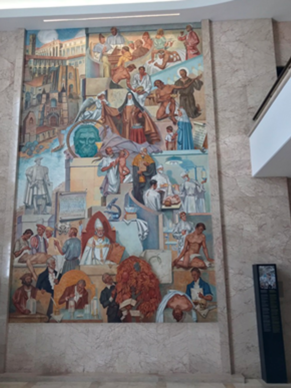 A tall wall mural depicting multiple historical scenes and figures arranged in stacked panels, with architectural backdrops, books, tools and symbolic objects woven throughout. The artwork features layered storytelling across different eras, framed by a marble wall with a display panel beside it.