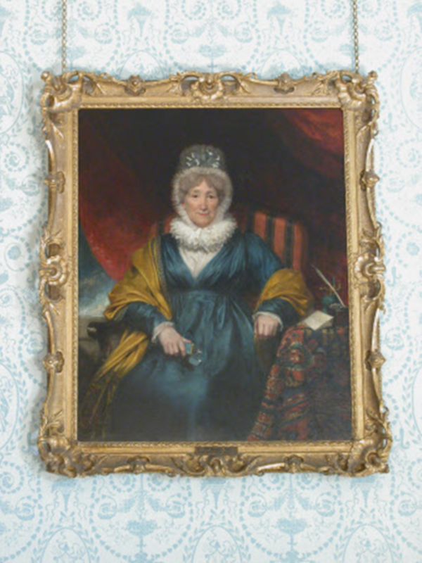 A framed oil portrait of Hannah More seated indoors, wearing a blue dress with a light shawl, shown against rich drapery and furnishings, in an ornate gilt frame on patterned wallpaper.