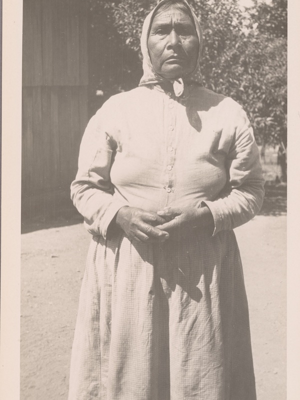 Full‑length black‑and‑white portrait of a person standing outdoors in a long, buttoned dress and head covering, facing the camera, with hands folded in front.