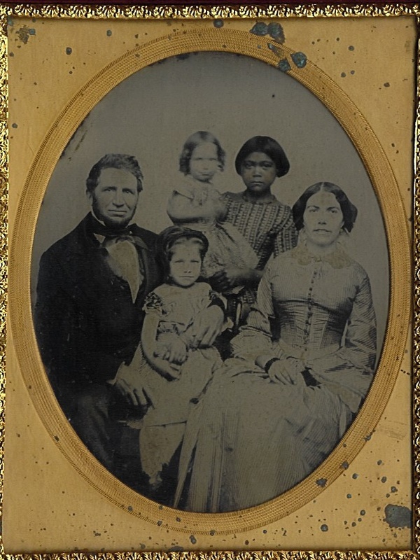 Oval‑framed black‑and‑white portrait of a group of adults and children posed indoors, seated and standing closely together in formal mid‑19th‑century clothing, facing the camera.
