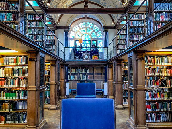 Interior of a historic library with tall wooden bookshelves filled with books, ornate vaulted ceiling with decorative patterns, a large arched window at the back, and a blue upholstered chair in the centre on a parquet floor.