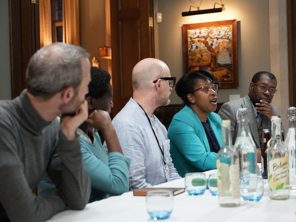 Group of people seated around a table in a meeting room, with water bottles and glasses on the table. The background shows a wooden door and a framed painting on the wall.
