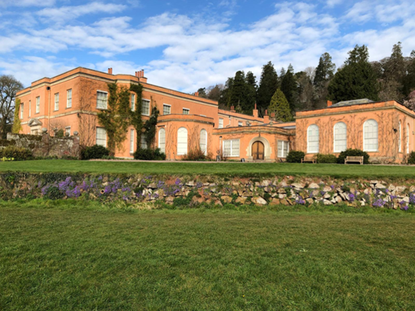 A wide view of a peach‑coloured historic building with arched windows and ivy‑covered walls, set behind a green lawn and low stone wall, with trees and a blue, partly cloudy sky in the background.