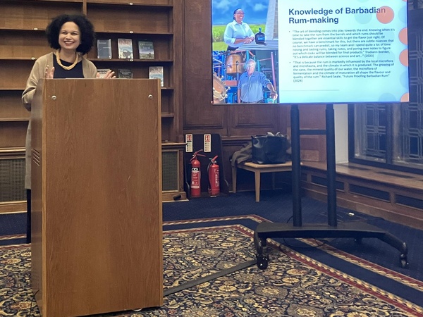 Dr. Tara Inniss speaking at a lectern during a presentation, with a slide on Barbadian rum-making displayed on a screen beside her.