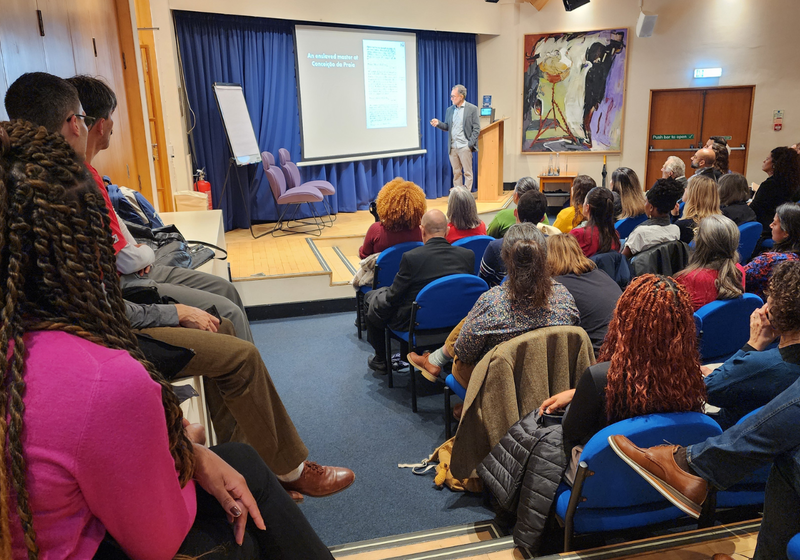 Audience listen as Professor João José Reis presents the lecture from a small stage in front of a powerpoint slide