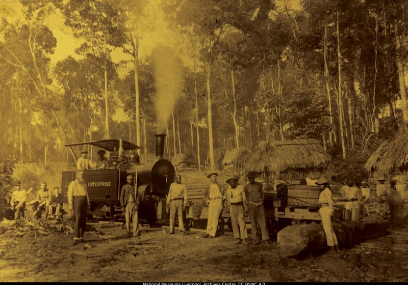 Archival image of a steam locomotive labeled “Enterprise” with smoke rising from its chimney, positioned on rail tracks in a forest clearing. Several workers stand around the train and timber logs, with thatched huts visible among tall trees in the background.