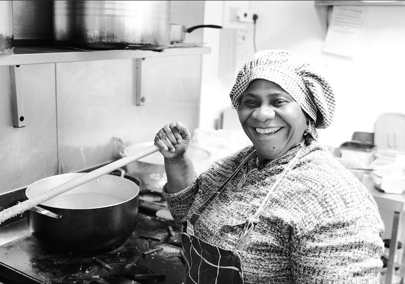 A person working in a kitchen stands beside a stove, stirring a large pot with a long wooden utensil. The setting includes metal cookware, a tiled wall, and various kitchen equipment in the background.