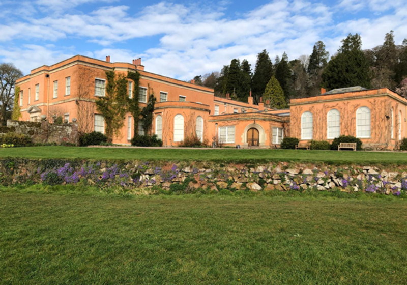 A wide view of a peach‑coloured historic building with arched windows and ivy‑covered walls, set behind a green lawn and low stone wall, with trees and a blue, partly cloudy sky in the background.