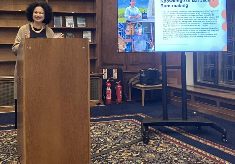 Dr. Tara Inniss speaking at a lectern during a presentation, with a slide on Barbadian rum-making displayed on a screen beside her.